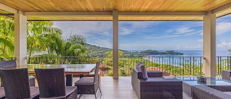 An outdoor patio with wicker furniture, a glass table, and a view of the sea and hills.
