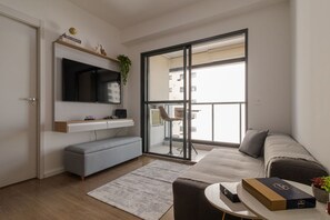 Uma sala de estar moderna com uma televisão de tela plana, um sofá cinza, uma mesa de centro e uma porta de vidro deslizante que dá para uma varanda externa.