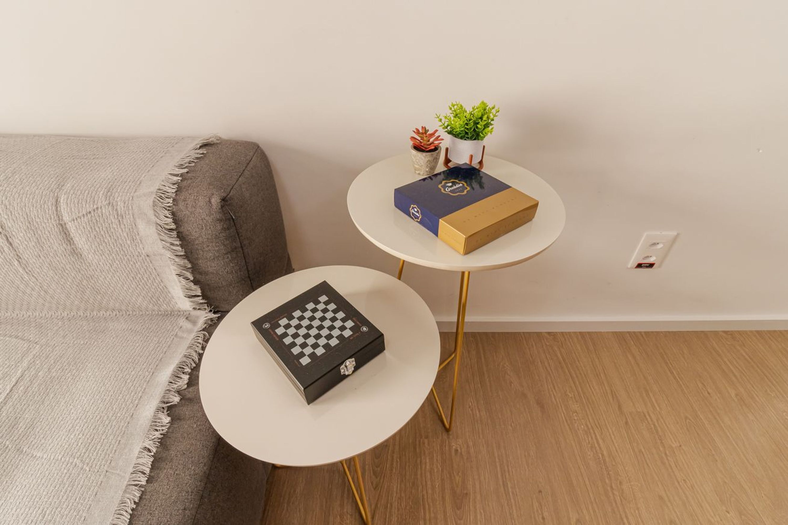 Um quarto com um sofá cinzento, uma pequena mesa redonda branca com um tabuleiro de xadrez e uma mesa redonda branca maior com uma caixa e uma planta.