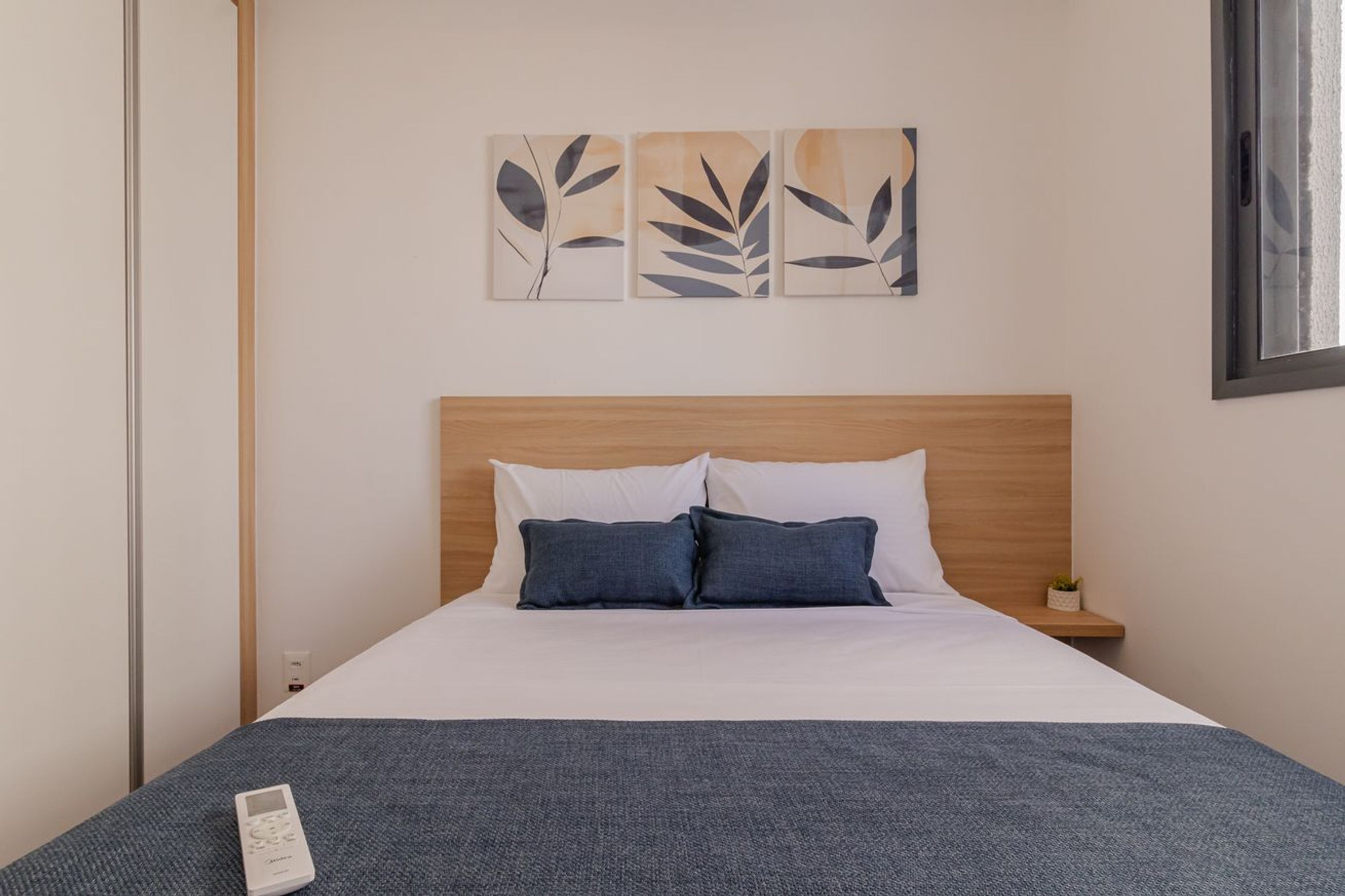 Quarto de hotel com cama, almofadas, mesa de cabeceira, controle remoto e obras de arte na parede.