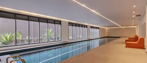 A swimming pool with a view of greenery outside, a tiled floor, and orange seating areas.