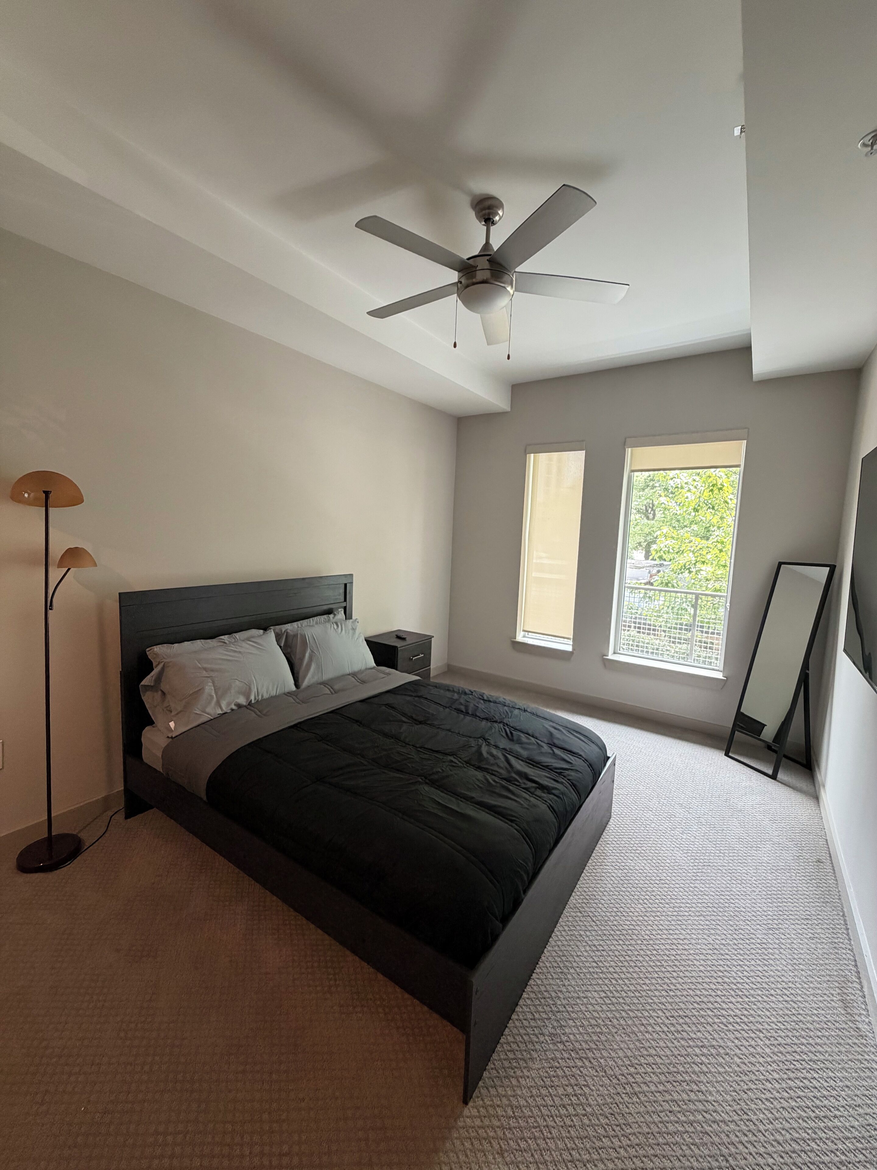 A bedroom with a bed, a ceiling fan, a nightstand, a floor lamp, and a window.