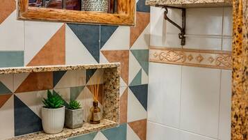Ein Badezimmer mit einem geometrischen Fliesenmuster, einer Toilette, einem Fenster mit Holzrahmen sowie einem Regal mit Pflanzen und einer Kerze.