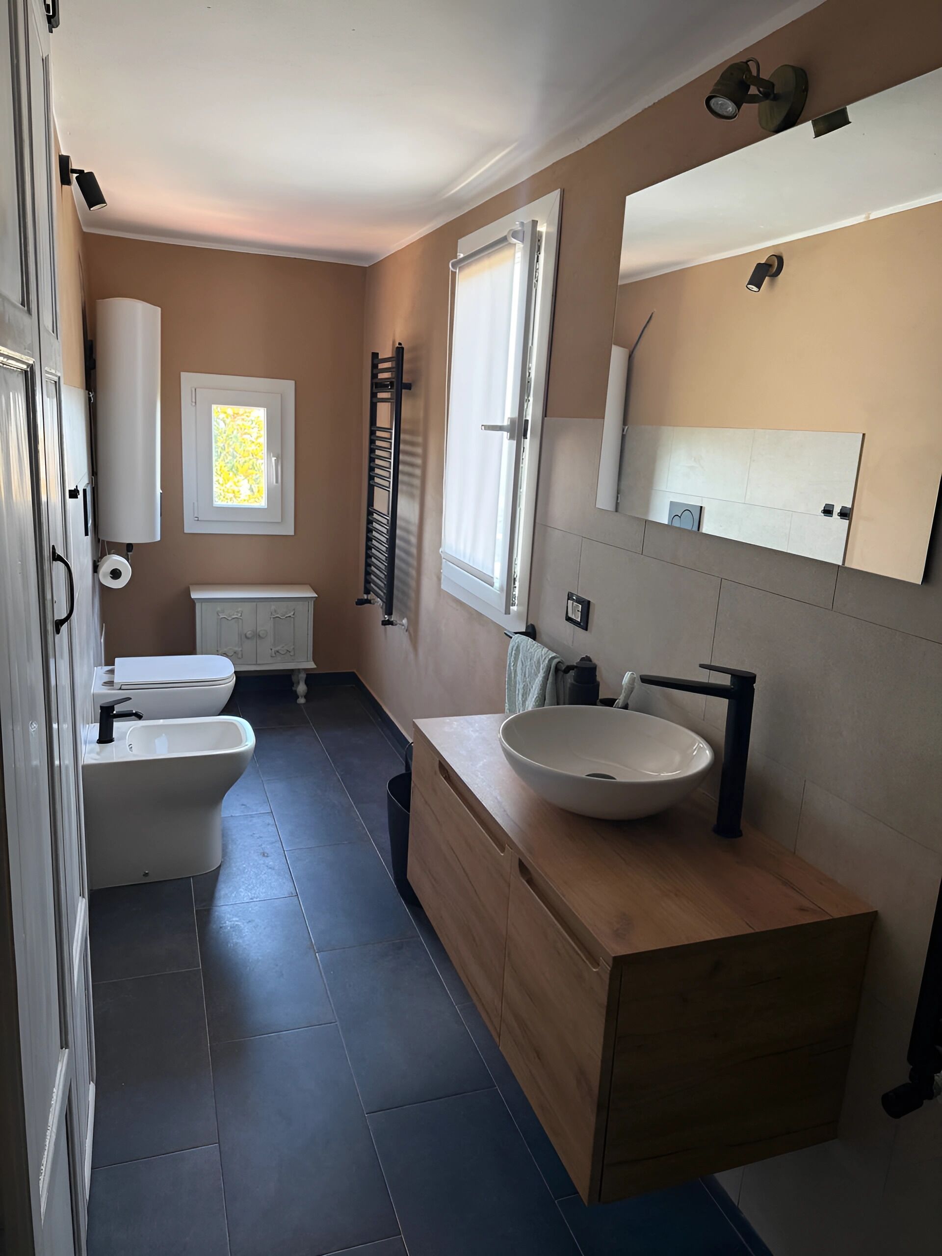A modern bathroom with a toilet, bidet, and sink. There is a window with a view of the outside, a towel rack, and a mirror above the sink.