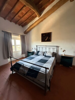Un dormitori amb un llit, bigues de sostre de fusta, una finestra amb cortines, una tauleta de nit amb una làmpada i un quadre penjat a la paret.