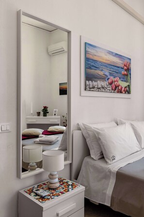 A bedroom with a bed, a bedside table with a lamp, a mirror, a painting, and a wall-mounted air conditioner. - Casa Ninetta - Terrace near Central Station (Palermo)