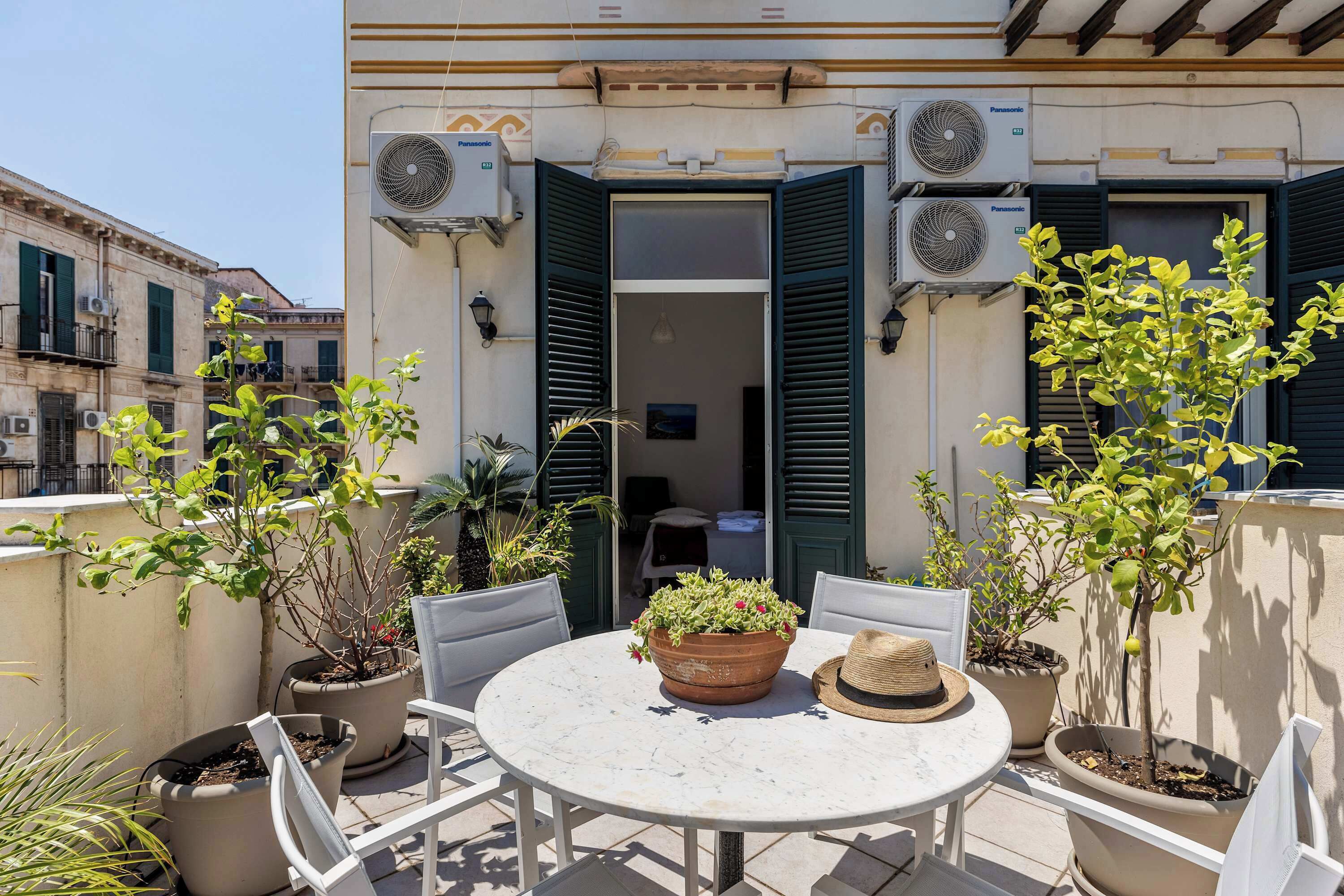 Un espace extérieur aménagé avec une table ronde et des chaises, entouré de plantes en pot et de climatiseurs.