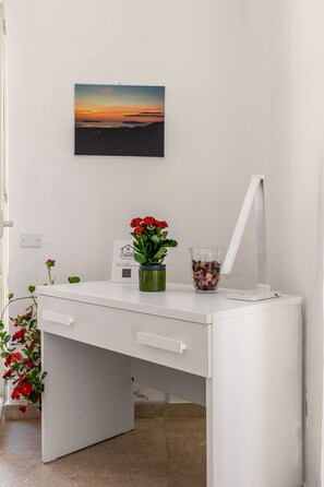Un bureau blanc avec un écran d’ordinateur, un vase rempli de fleurs rouges et, au mur, un tableau représentant un coucher de soleil.