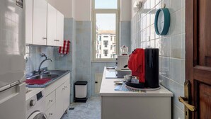 A compact kitchen with white cabinets, a sink, a coffee machine, and a window providing a view of buildings outside. - Casa Ninetta - Terrace near Central Station (Palermo)