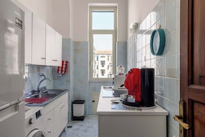 Eine kompakte Küche mit weißen Schränken, einer Spüle, einer Kaffeemaschine und einem Fenster mit Blick auf Gebäude.