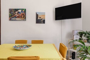 A dining area with a table set for four, a television mounted on the wall, and a shelf with plants and a lamp. - Casa Ninetta - Terrace near Central Station (Palermo)
