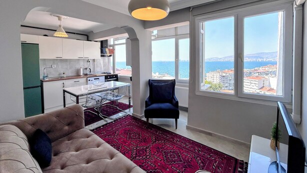 A modern living room with a sofa, a dining table with chairs, a red patterned rug, and a view of the sea through large windows. - Renovated comfy apartment with unique sea view (Konak)