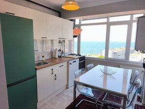 Une cuisine moderne avec vue sur la mer, un coin repas doté d’une table en verre et un réfrigérateur vert.