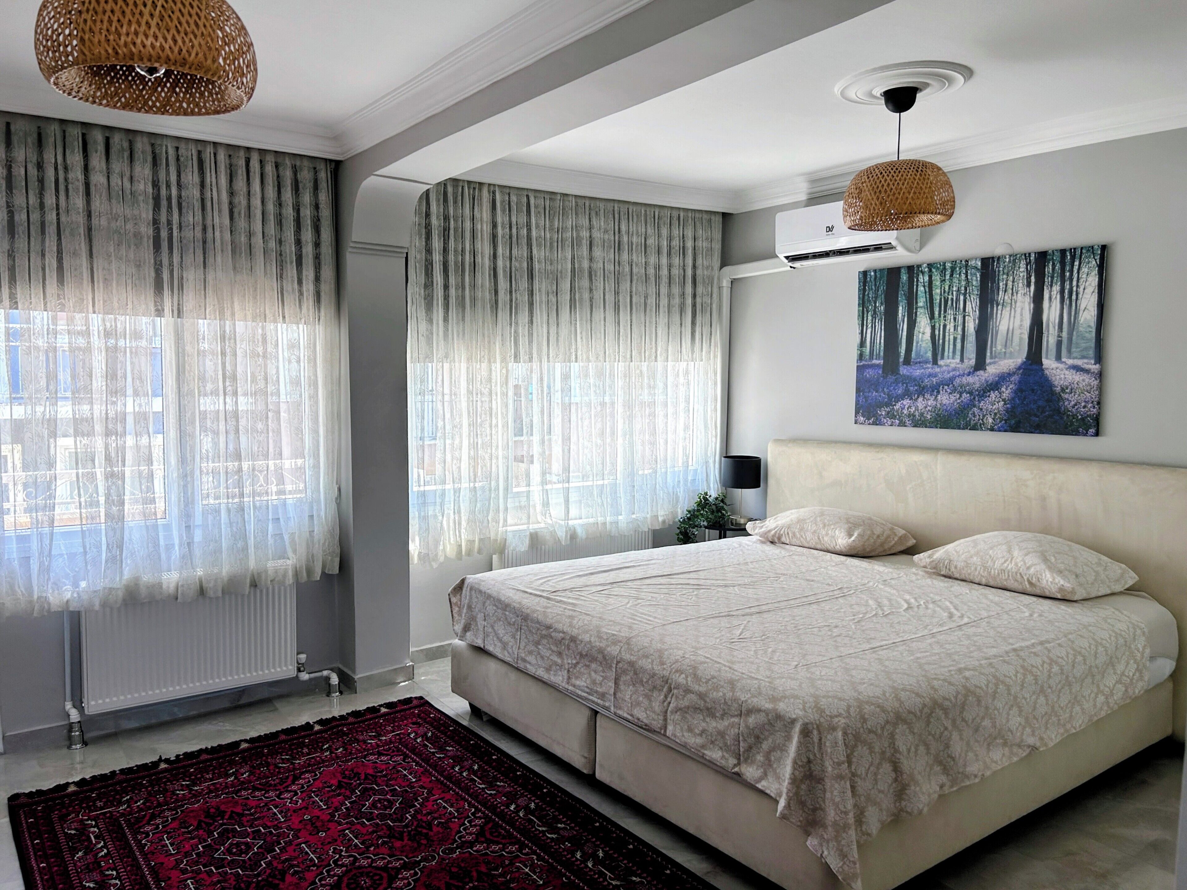 A bedroom with a bed, a radiator, a window with sheer curtains, a painting on the wall, and a hanging light fixture.