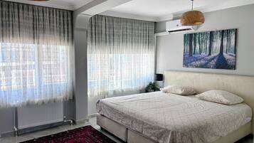 A bedroom with a bed, a radiator, a window with sheer curtains, a painting on the wall, and a hanging light fixture.