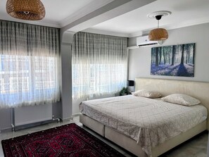 Un dormitorio con cama, un radiador, una ventana con cortinas traslúcidas, un cuadro en la pared y una lámpara colgante.