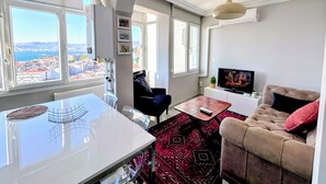 A modern living room with a sofa, a TV, a dining table, and large windows offering a city view. - Renovated comfy apartment with unique sea view (Konak)
