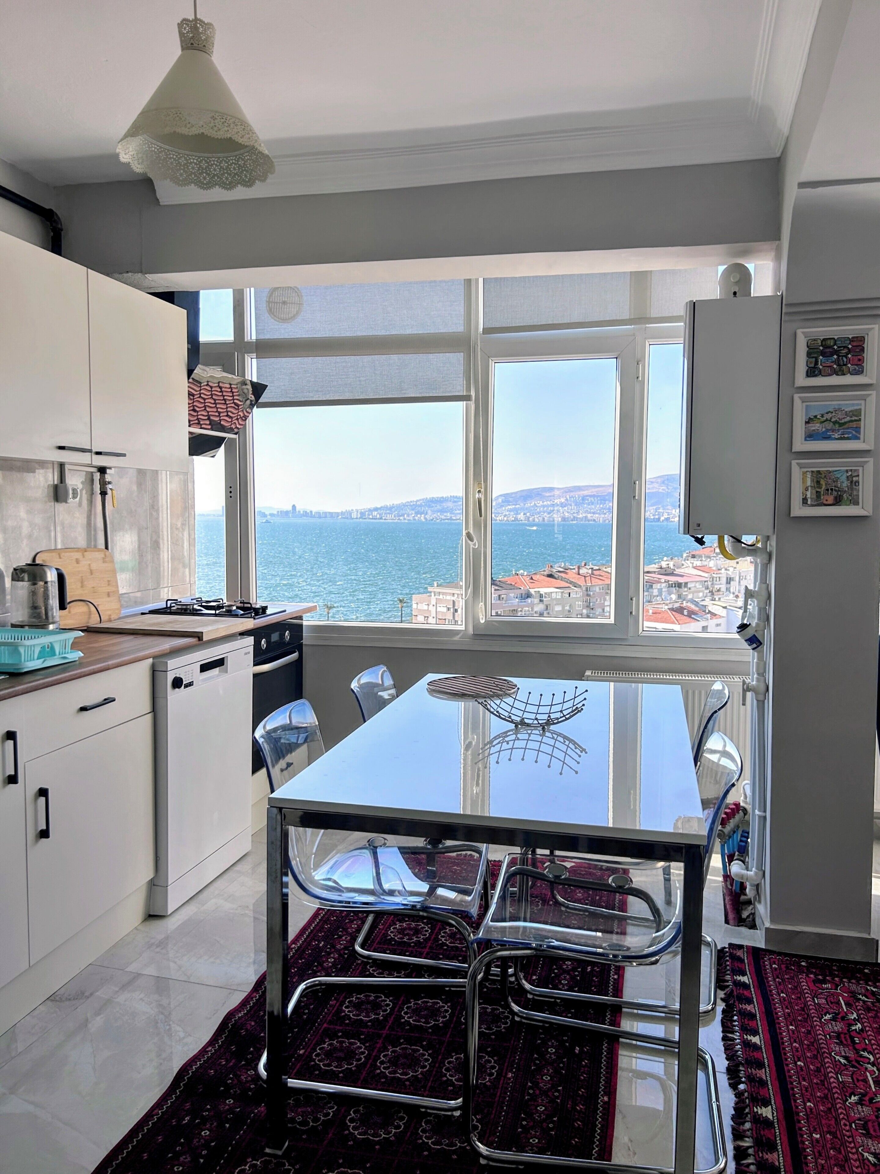 A kitchen with a view of the sea, a dining table with chairs, and a red patterned rug.