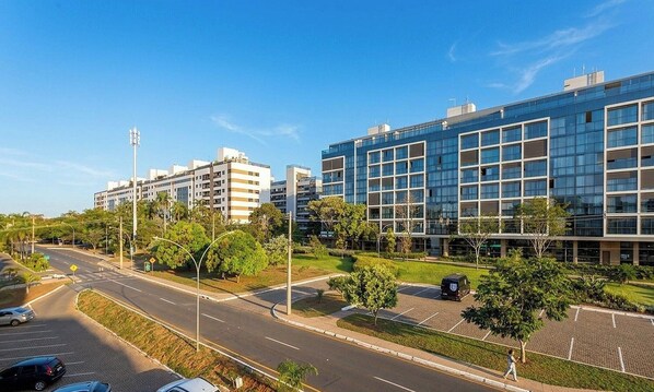A modern building with a glass facade, surrounded by a well-maintained park and a road with parked cars. - Studio in Condo. w/ AC in Setor Noroeste Pzs0000 (Brasília)