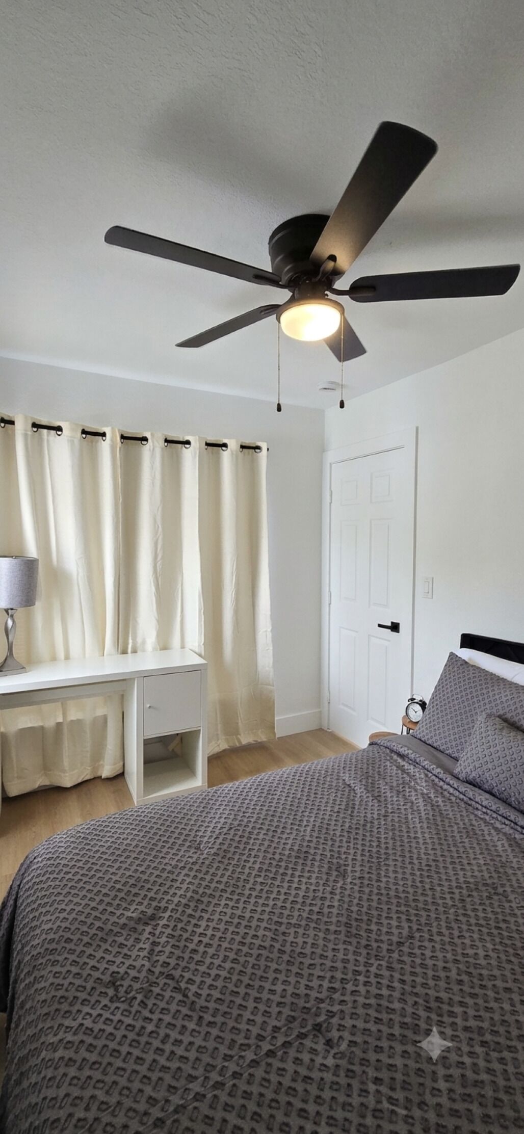 A bedroom with a bed, a desk, a chair, a ceiling fan, and a door.