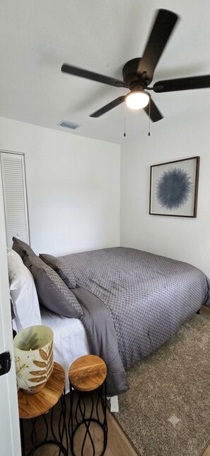 Een slaapkamer met een bed, een plafondventilator, een kunstwerk in een lijst en een klein tafeltje met een kop.