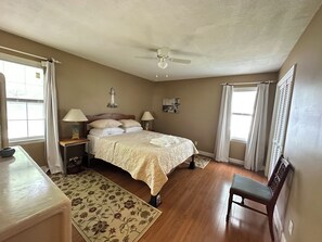Ein Schlafzimmer mit einem Bett, zwei Nachttischen, einem Hocker, einem Sessel, einem Deckenventilator und einem Fenster mit Vorhängen.