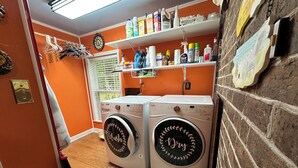 Uma lavanderia com duas máquinas de lavar de abertura frontal, uma prateleira com produtos de limpeza e uma janela com persianas.