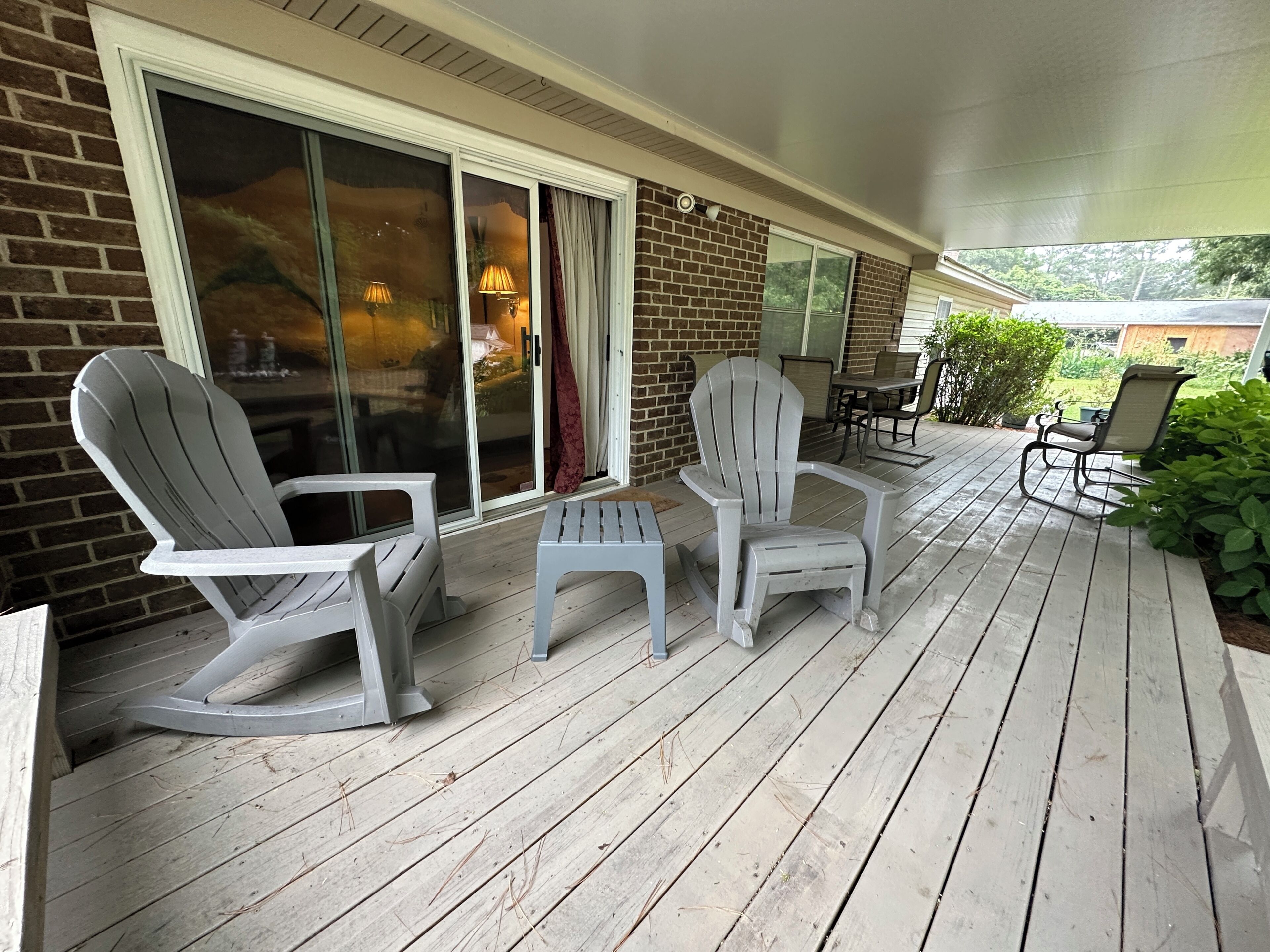 Un espace extérieur aménagé, abrité, avec deux fauteuils Adirondack et une petite table, un mur en briques et une porte-fenêtre coulissante donnant sur un patio.