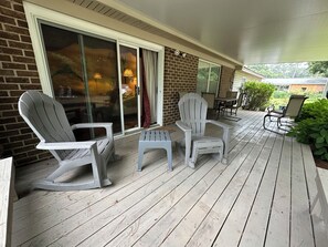 A covered outdoor seating area with two Adirondack chairs and a small table, a brick wall, and a sliding glass door leading to a patio.
