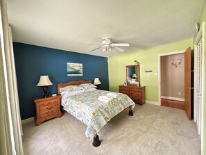 Une chambre à coucher avec un lit, deux tables de chevet, un ventilateur de plafond, une commode et une porte.