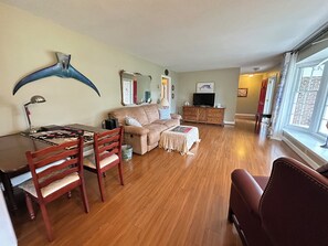 Un salon avec un plancher en bois, un canapé, une table à manger avec des chaises, un téléviseur posé sur un meuble en bois et une grande fenêtre avec des rideaux.