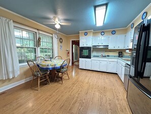 A kitchen with wooden flooring, white cabinets, a dining table with chairs, a ceiling fan, and a window with curtains. - 3BR Coastal Escape with Bogue Sound Access (Morehead City)