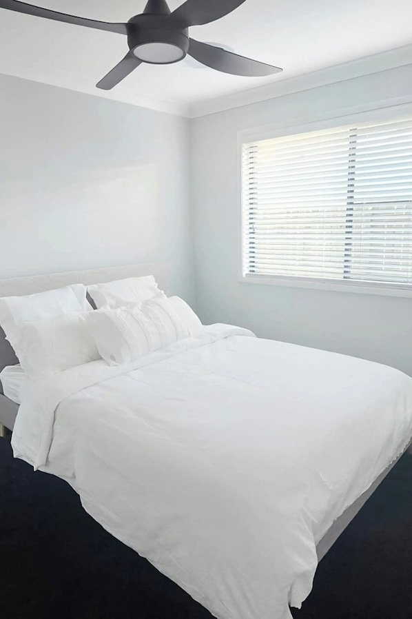 A bedroom with a bed, white bedding, a ceiling fan, and a window with blinds.