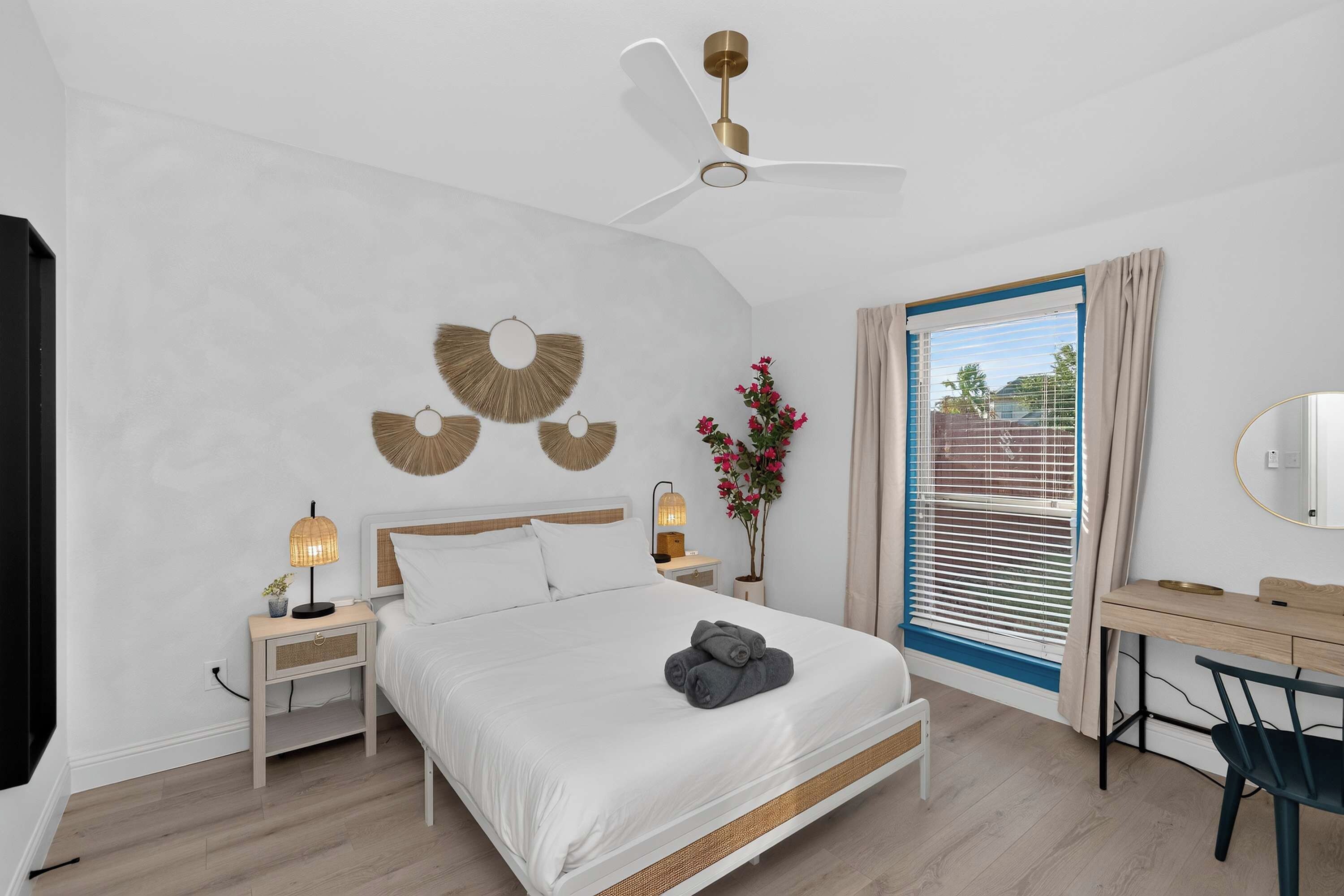 Une chambre moderne comprenant un lit, des tables de chevet, un bureau, une chaise, un ventilateur de plafond et une fenêtre avec des rideaux.