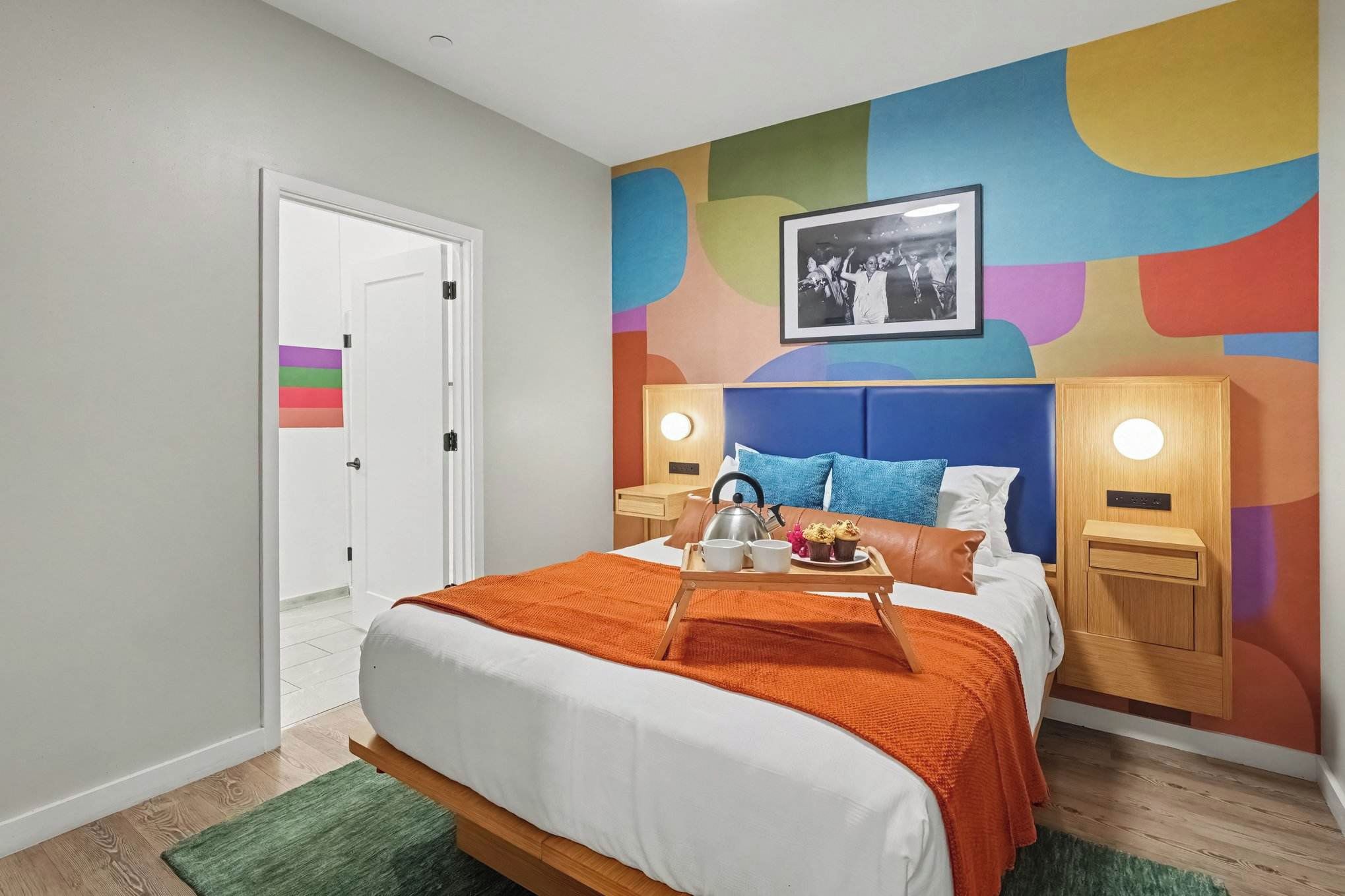 Una habitación de hotel moderna con una cama, una mesita de noche con una tetera, una bandeja con tazas y un hervidor, un llamativo mural abstracto en la pared y una puerta con una bandera arcoíris.