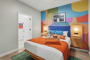 Una habitación de hotel moderna con una cama, una mesita de noche con una tetera, una bandeja con tazas y un hervidor, un llamativo mural abstracto en la pared y una puerta con una bandera arcoíris.