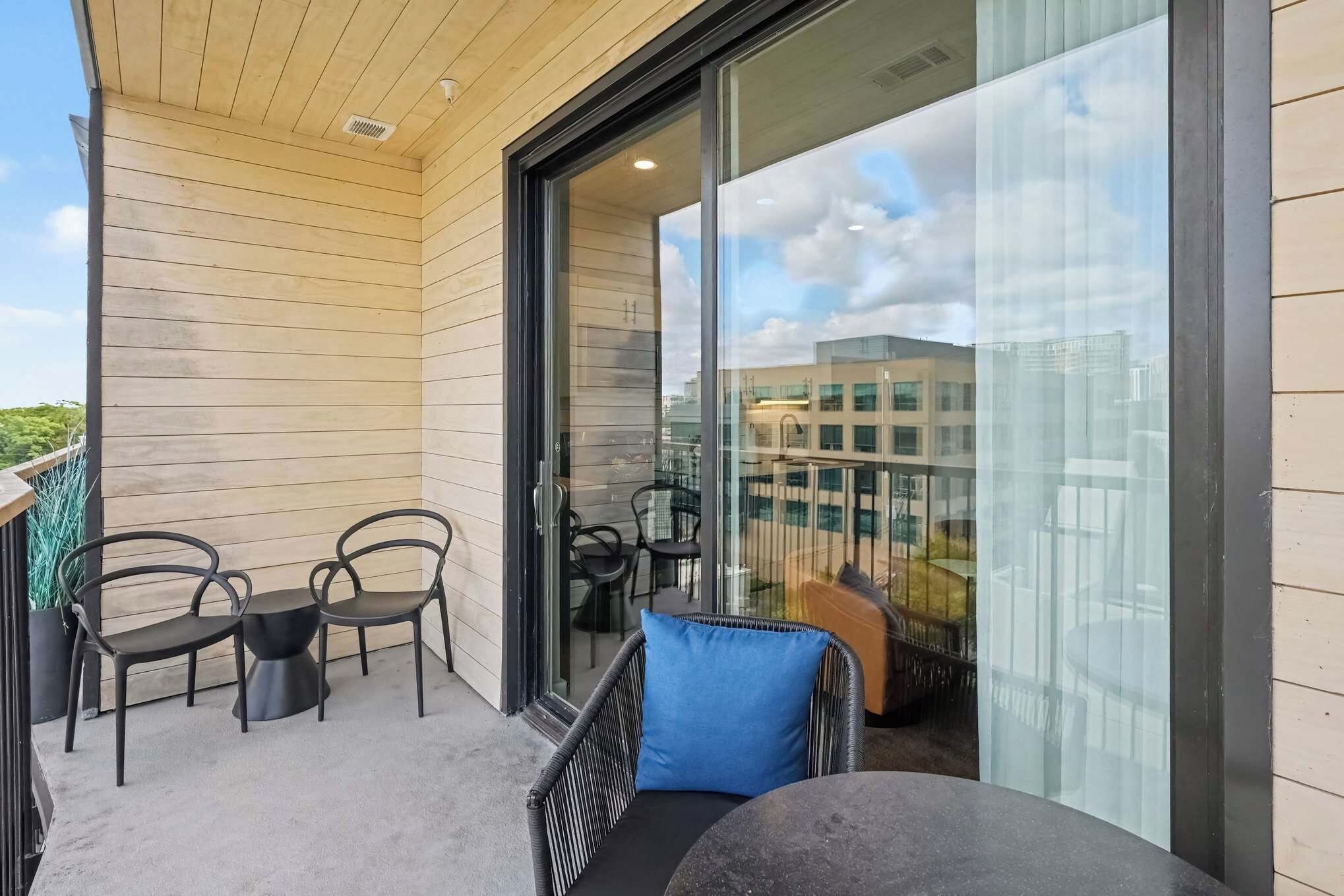 A balcony with a wooden floor, a table, two chairs, a blue cushion, and a glass door leading to an indoor area with chairs and a sofa.