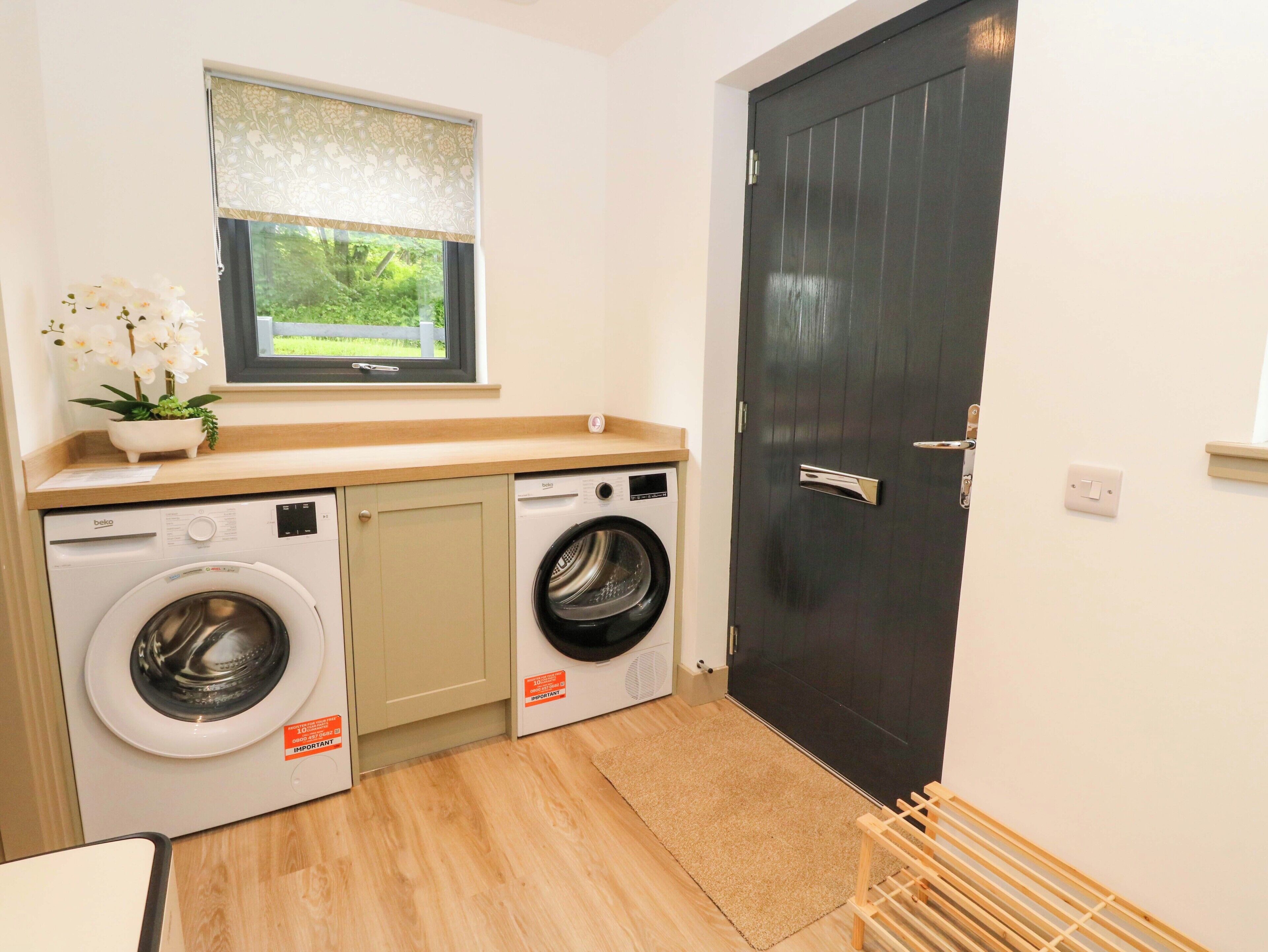 A laundry room with two front-loading washing machines, a small window with a view of greenery, and a dark door.