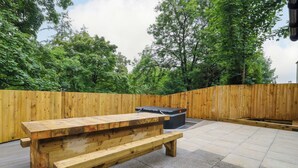 An outdoor wooden bench area with a stone floor, surrounded by a wooden fence and trees. - Nevis Bridge House (Fort William)