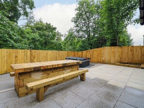 An outdoor wooden bench area with a stone floor, surrounded by a wooden fence and trees. - Nevis Bridge House (Fort William)