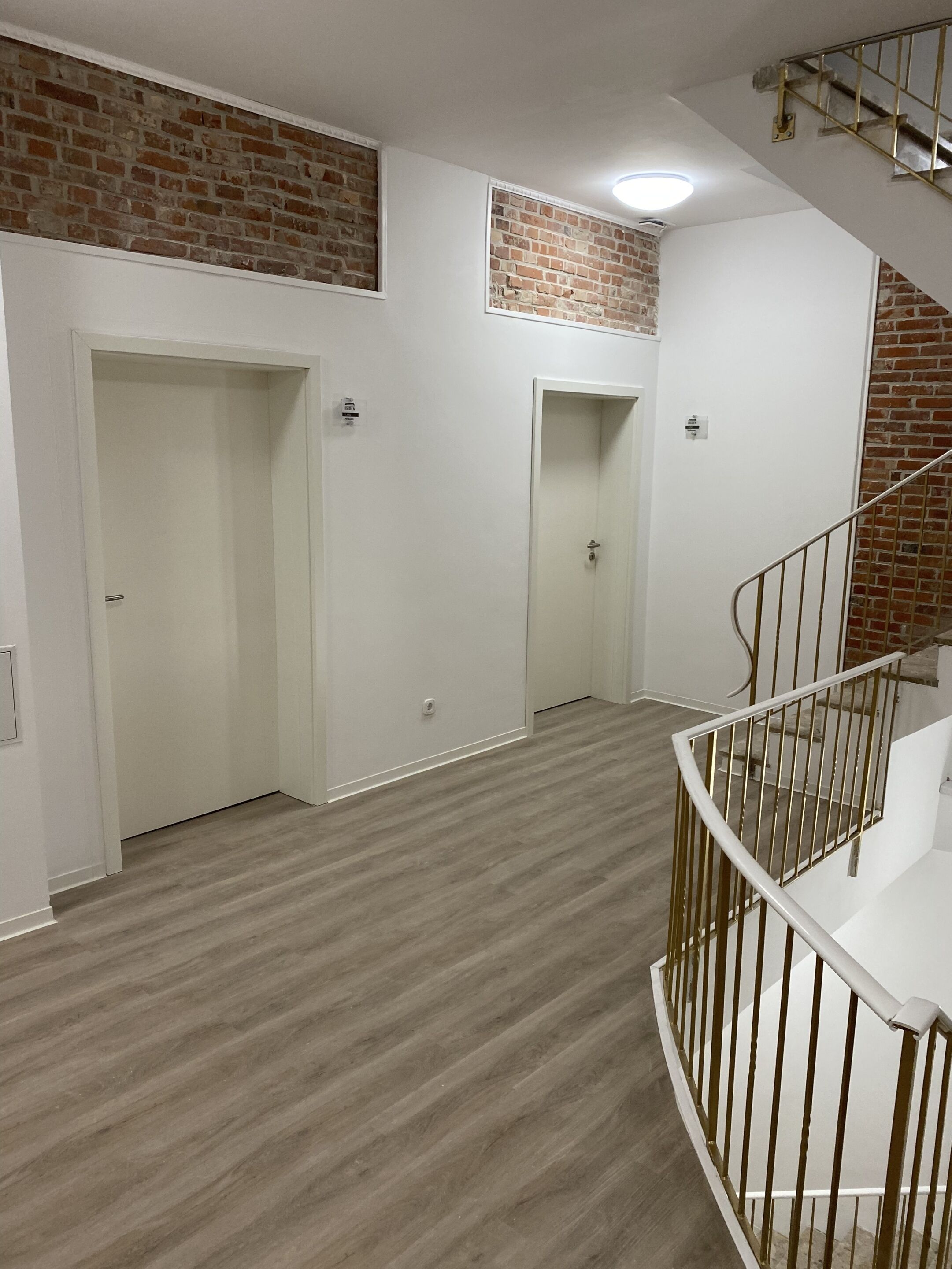 Ein mehrgeschossiges Interieur mit Holzboden, weißen Wänden und einer Ziegelwand als Blickfang. Eine Treppe mit Metallgeländer ist ebenfalls zu sehen.