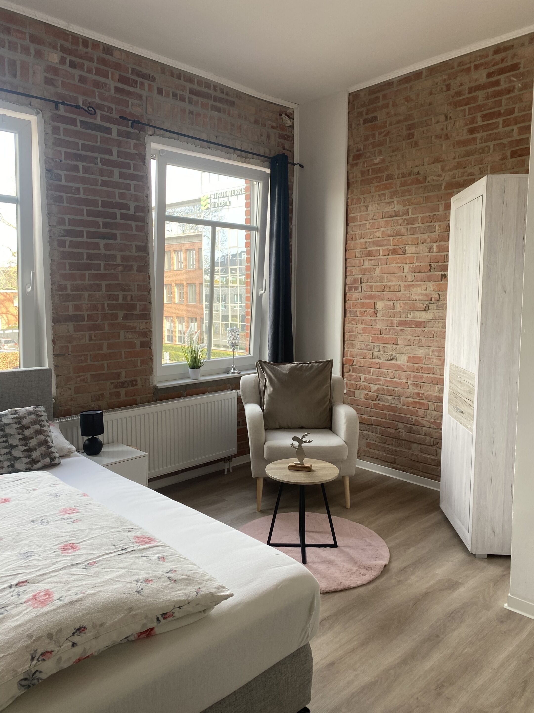 Ein Zimmer mit Backsteinwand, einem Bett mit floralem Bettzeug, einem weißen Sessel, einem kleinen runden Tisch und einem Heizkörper.