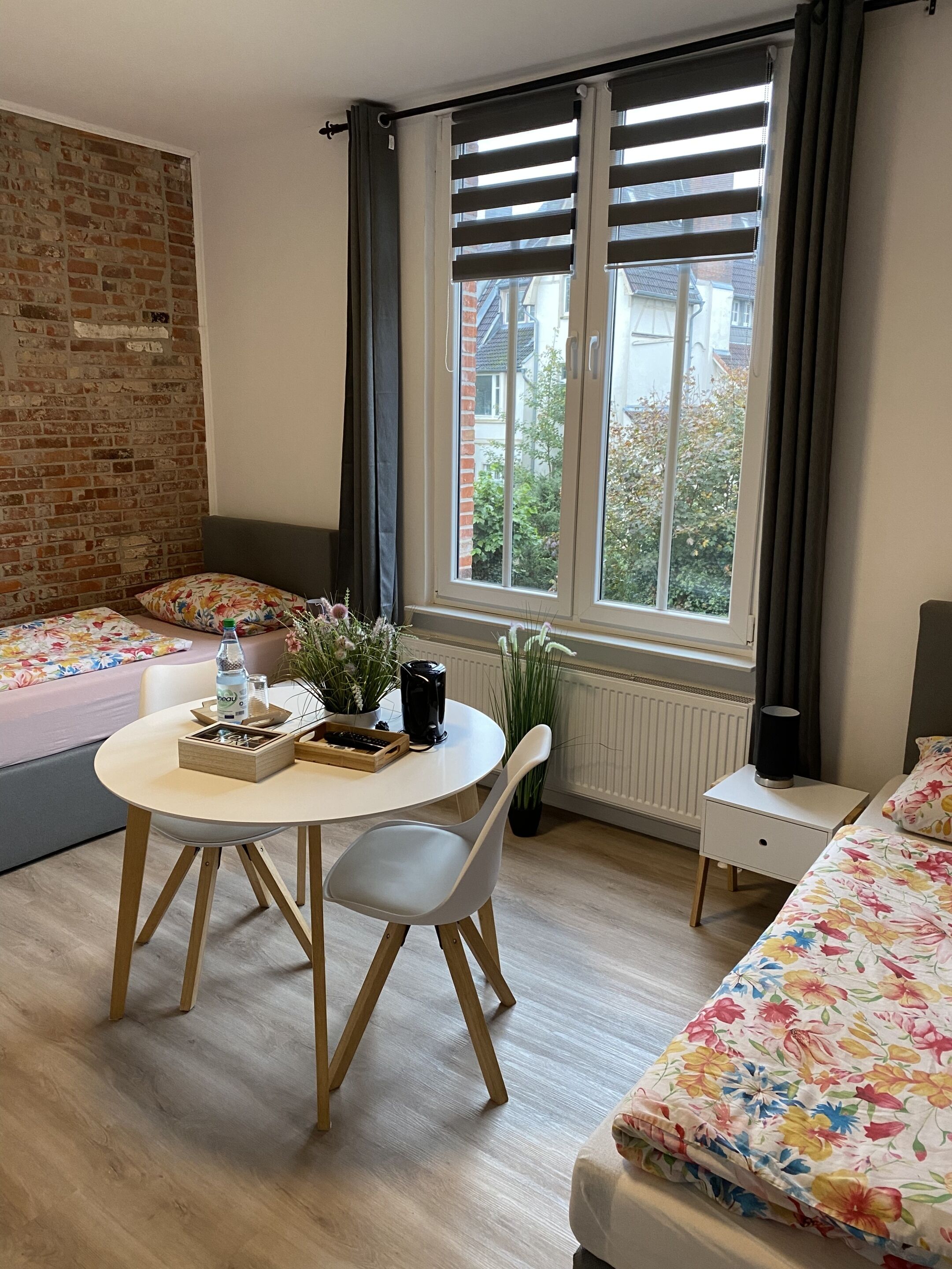 Een gezellige kamer met een bakstenen muur, een raam met verstelbare zonwering, een ronde tafel met een stoel, een bed met bloemenprint beddengoed en een radiator.