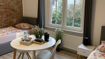 Een gezellige kamer met een bakstenen muur, een raam met verstelbare zonwering, een ronde tafel met een stoel, een bed met bloemenprint beddengoed en een radiator.
