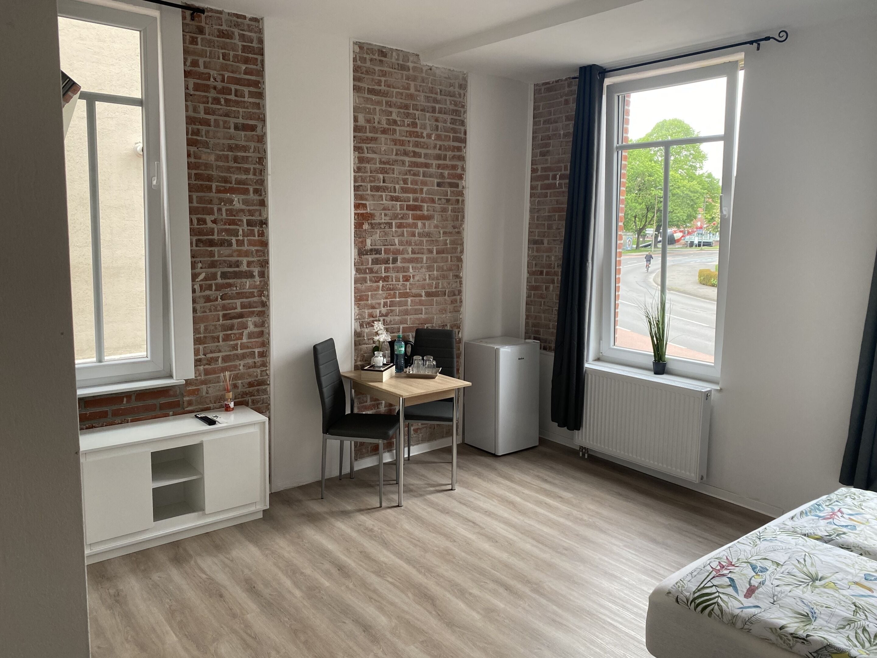 A room with a brick wall, a window with a view of the street, a radiator, a desk with a chair, and a bed with a patterned bedspread.