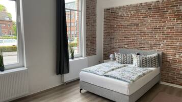 A bedroom with a brick wall, a bed with patterned bedding, a radiator, and a window with a curtain.