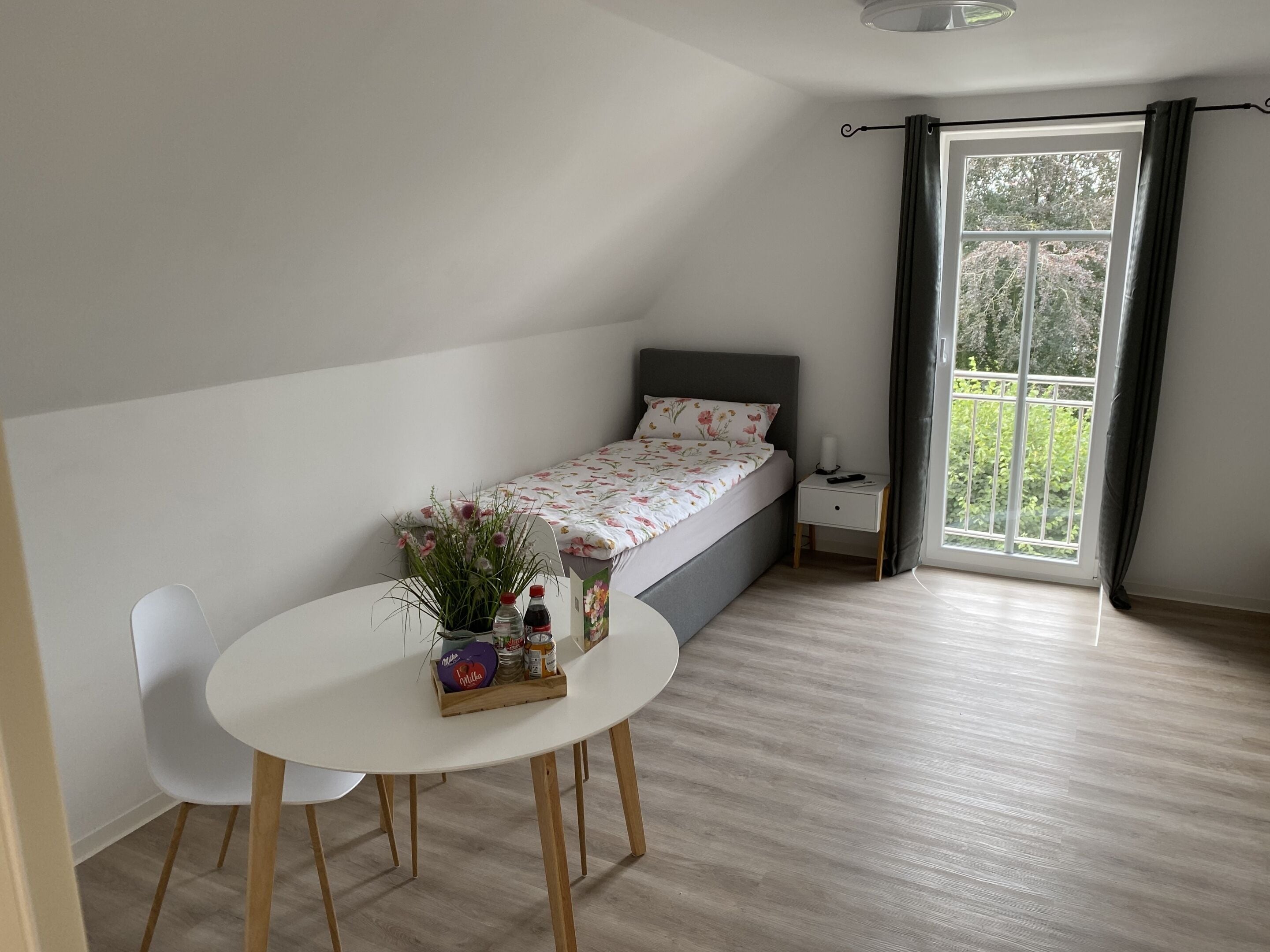 A small, single-bed room with a window, a bedside table, a chair, and a table with snacks.