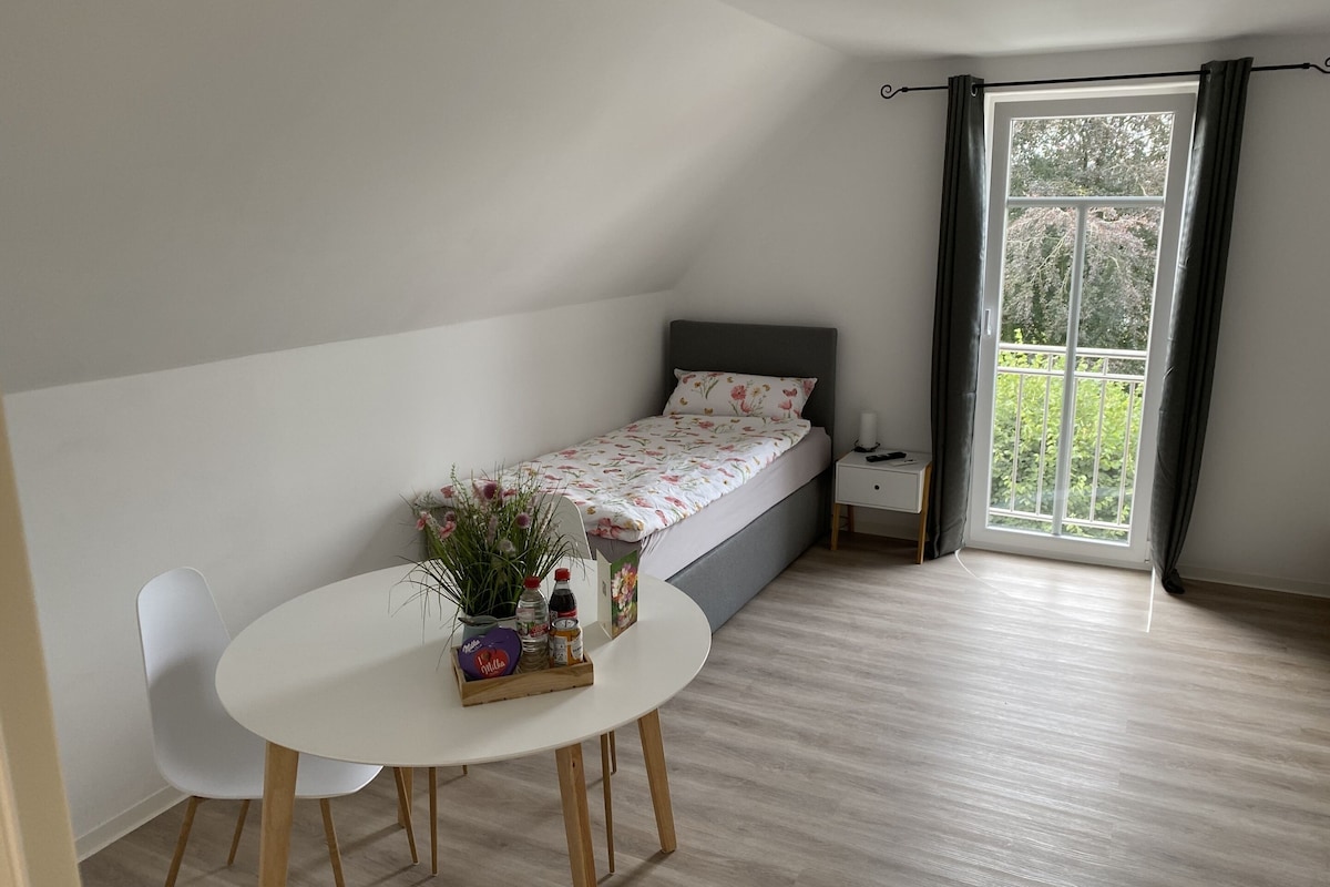 Quarto pequeno com uma cama de solteiro, janela, mesa de cabeceira, cadeira e mesa com lanches.