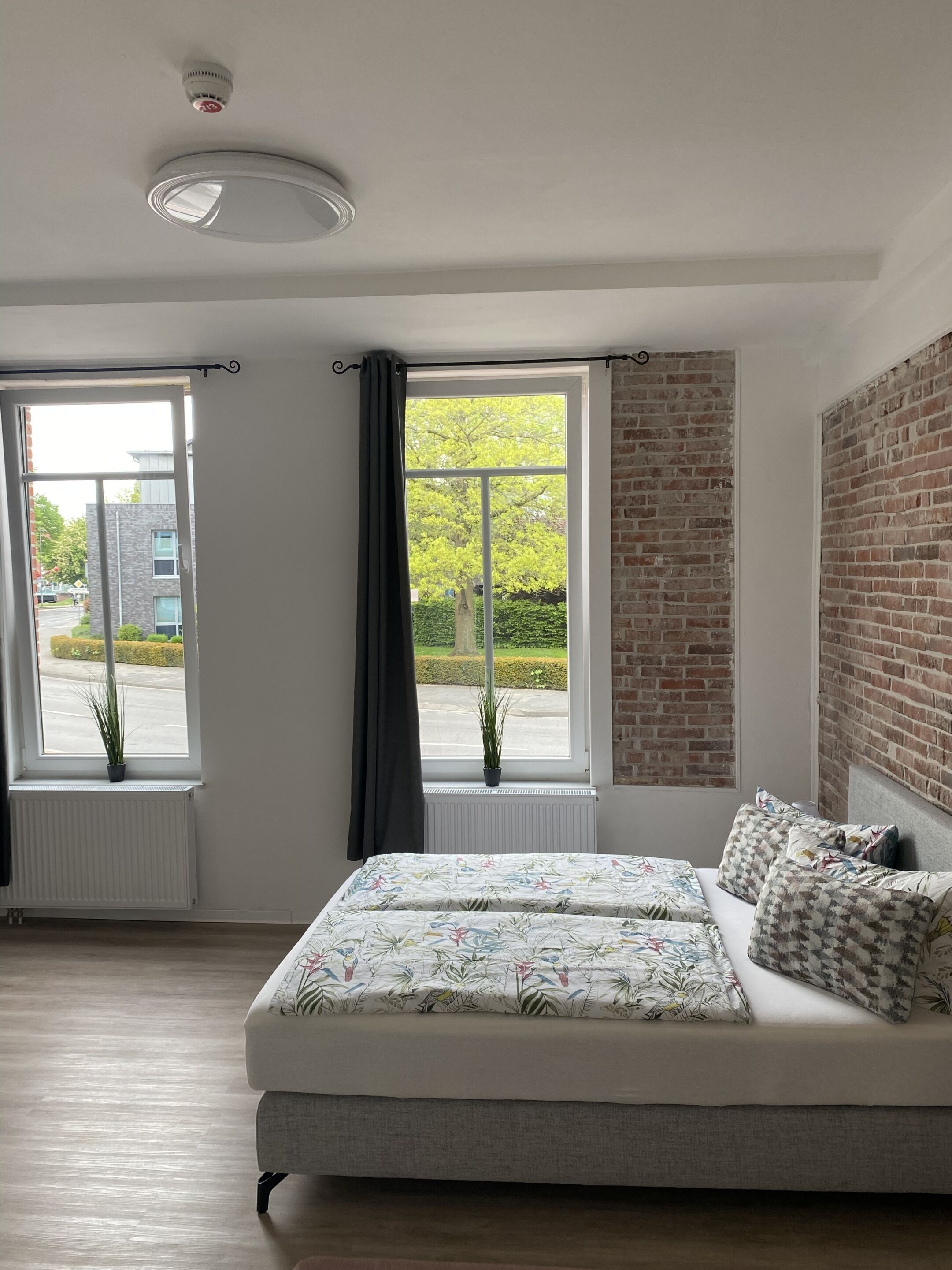 Un dormitorio con cama, dos ventanas, un radiador, una lámpara de techo y una pared de ladrillo.