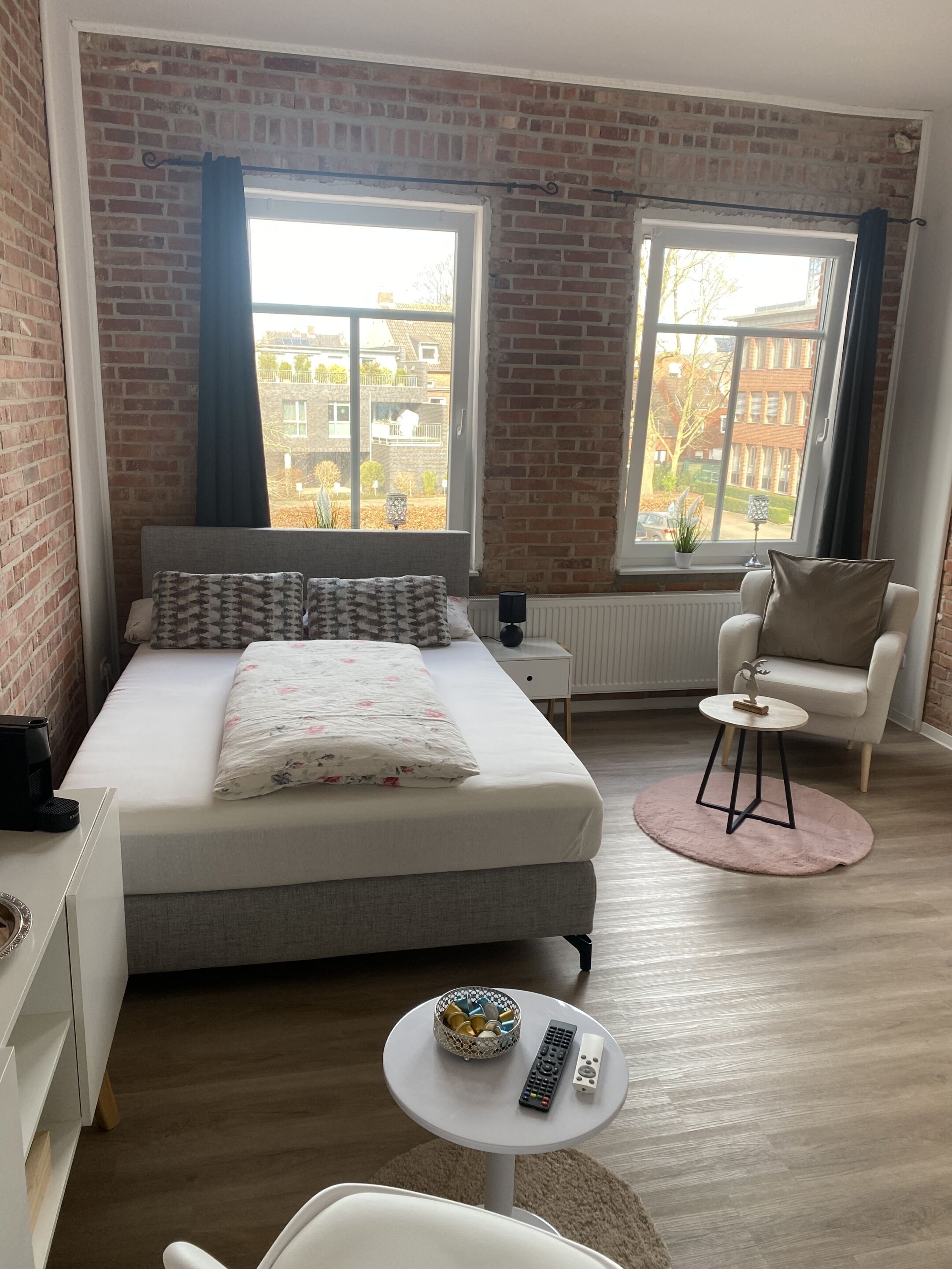 A modern bedroom with a large bed, two windows, a small round table with a basket and remote controls, and a white armchair.